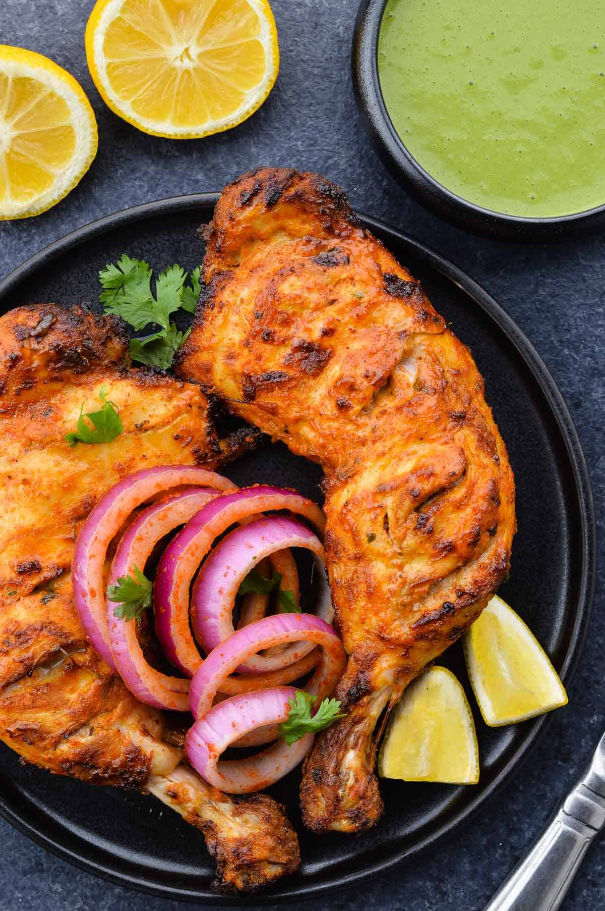 two tandoori chicken legs in a plate, along with laccha pyaaz, a couple lemon wedges & chopped coriander leaves, and a bowl of green chutney on the side.