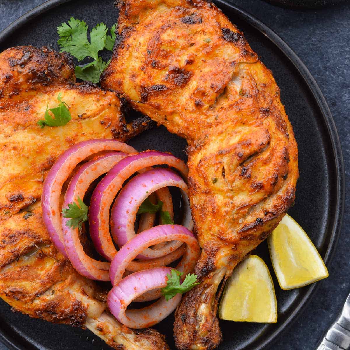 two tandoori chicken legs in a plate, along with laccha pyaaz, a couple lemon wedges & chopped coriander leaves.