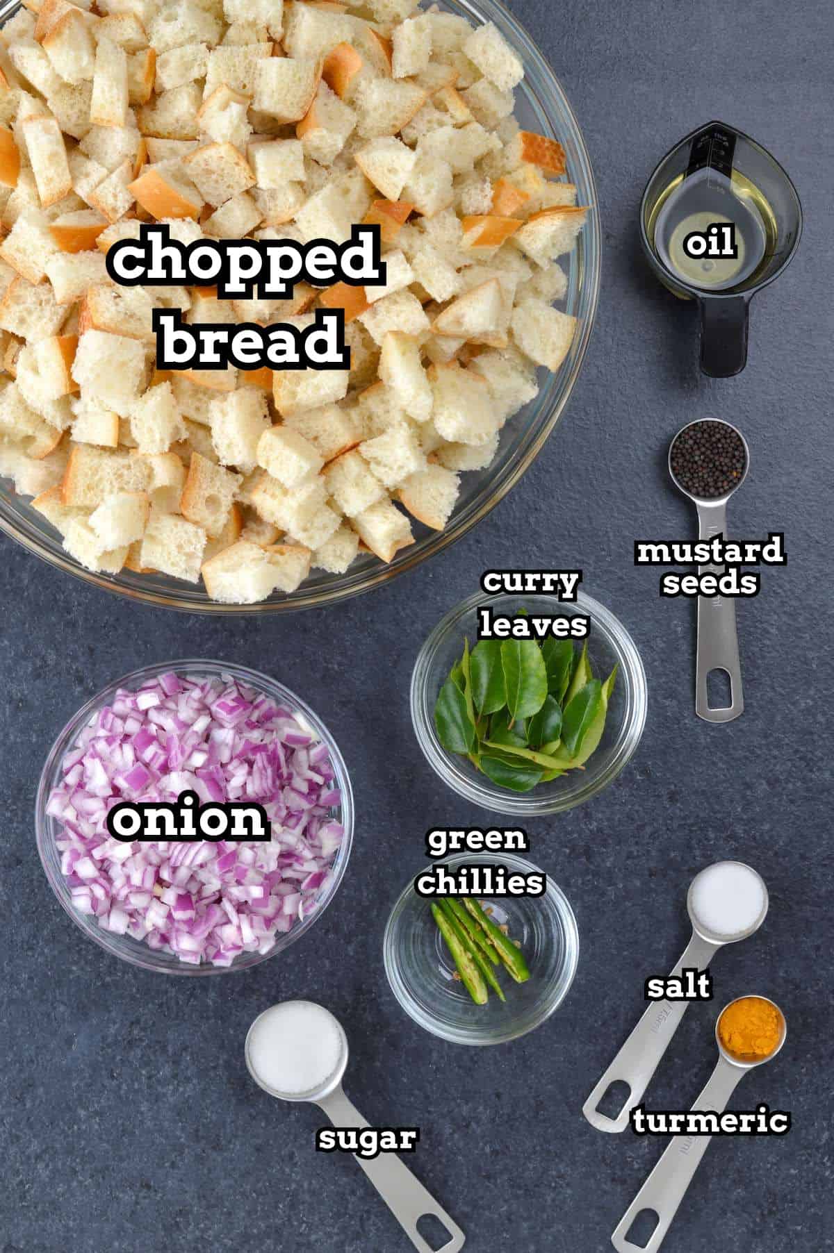 Top view of all the ingredients for making bread upkari, including chopped bread, onions, curry leaves, green chillies, and other seasonings, arranged in labeled bowls and spoons.