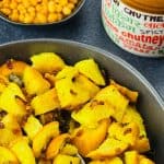 A bowl of bread upkari, with a spoon in it, served along with a small jar of Indian achar and a crunchy snack on the side.