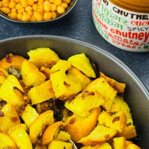 A bowl of bread upkari, with a spoon in it, served along with a small jar of Indian achar and a crunchy snack on the side.