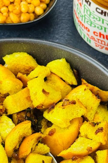 A bowl of bread upkari, with a spoon in it, served along with a small jar of Indian achar and a crunchy snack on the side.