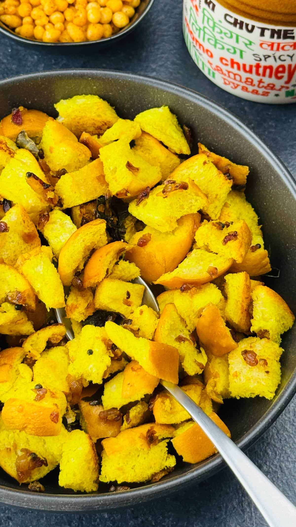 A bowl of bread upkari, with a spoon in it, served along with a small jar of achar and a crunchy snack on the side.